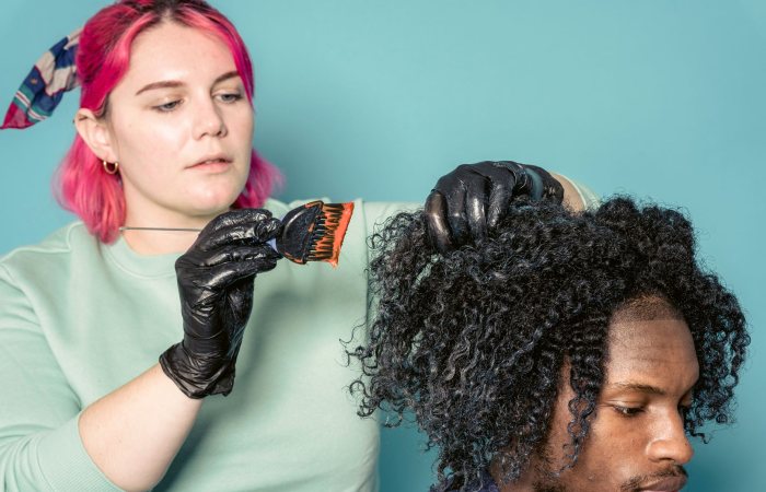 Serious woman with dyed hair applying paint on hair of African American model in studio against blue background